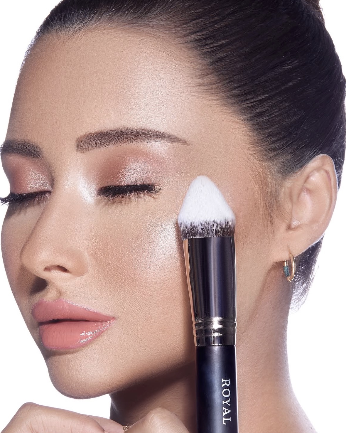 A close-up of luminous skin, softly shimmering eyes and pink lips with a makeup brush beside the cheek.