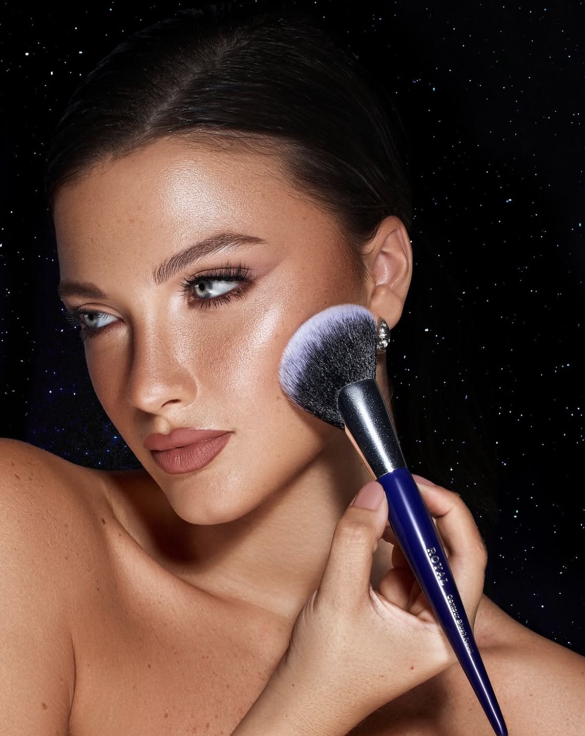 Warm glowing skin, softly defined eyes and a brown rose lip against a starry dark background.